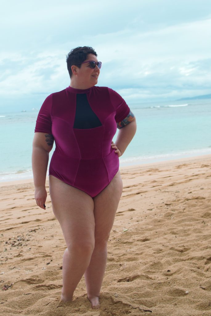 Front profile of model at beach. Wearing Chromat Tidal II lycra velvet one-piece swimsuit: Red-Violet, high neck, short sleeves, and a bikini-cut thigh. Triangular black mesh chest panel. Plus sized.