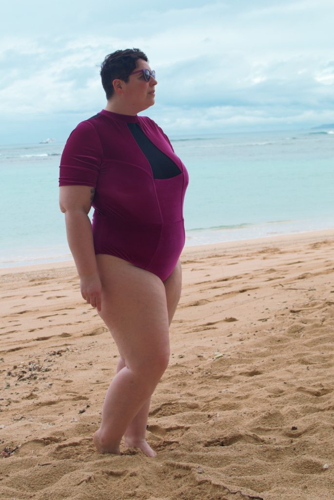 Side profile of model at beach. Wearing Chromat Tidal II lycra velvet one-piece swimsuit: Red-Violet, high neck, short sleeves, and a bikini-cut thigh. Triangular black mesh chest panel. Plus sized.