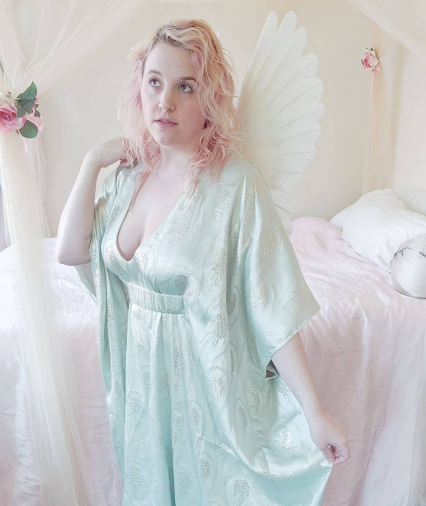 Photo is of a woman in soft blue silk caftan with angel wings on back standing in front of bed with tulle drapes and pink comforter.