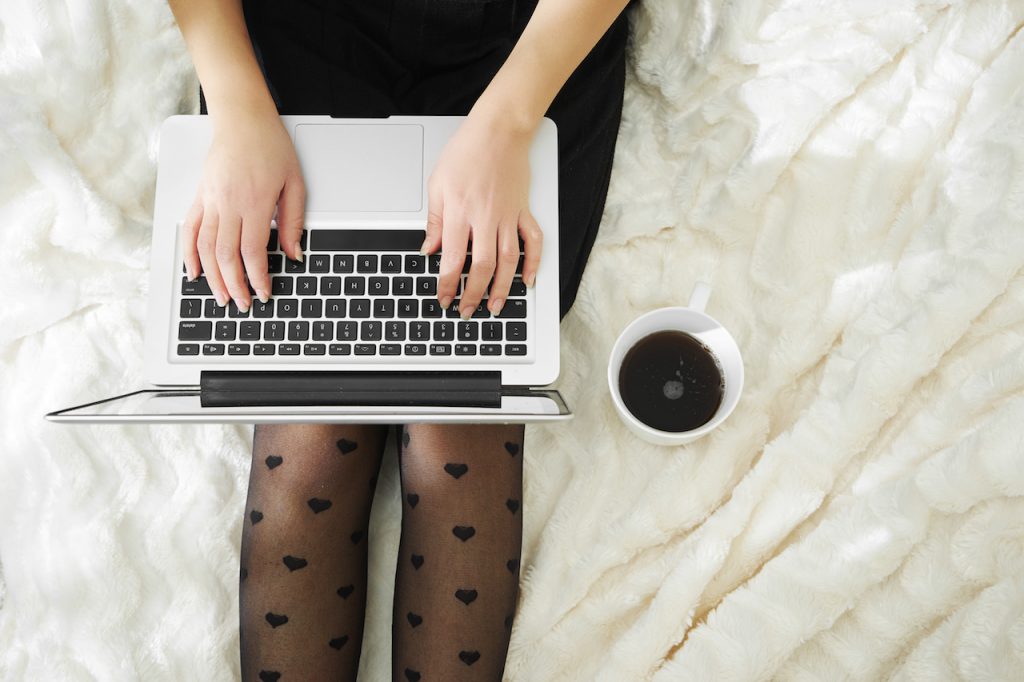 Laptop on the lower torso of a model sitting in bed typing. 