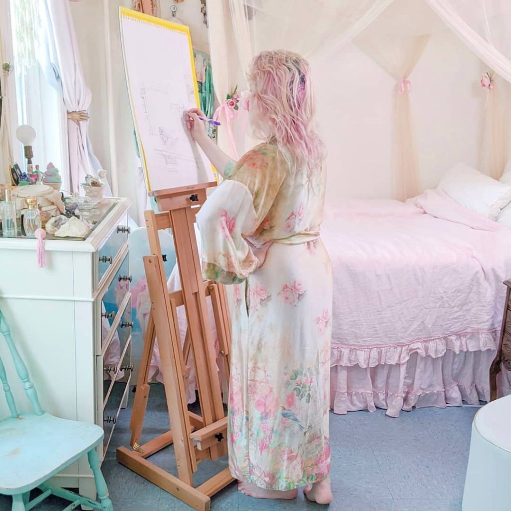 An example of silk lingerie you can use our oil removal technique on. Photo is of a woman with pink hair standing and drawing in front of easel in her bedroom. The bed has pink sheets, there's a mint green chair, and the dresser has perfume bottles and other accessories. The woman wears a light colored floral silk robe, yellow background with pink flowers and blue birds.