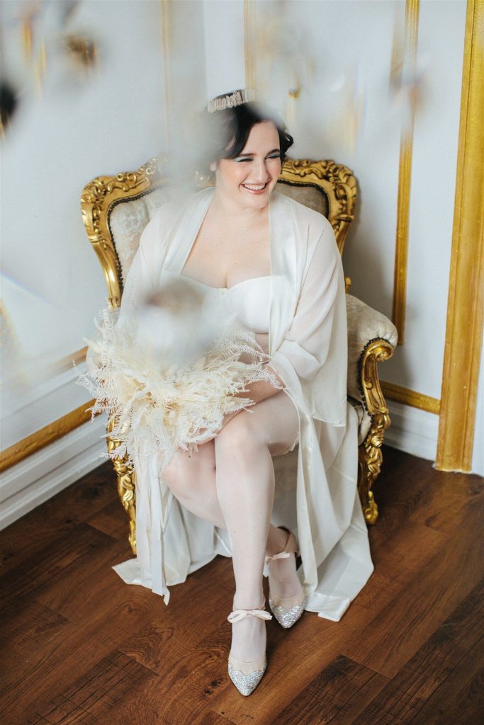 Model sitting wearing white, bridal lingerie set for larger busts. Curvy Kate, strapless Luxe Basque. Karolina Laskowska bespoke, lace button-back, ouvert knickers. Glamory Hosiery stockings. The Giving Bride Nora long robe. 
