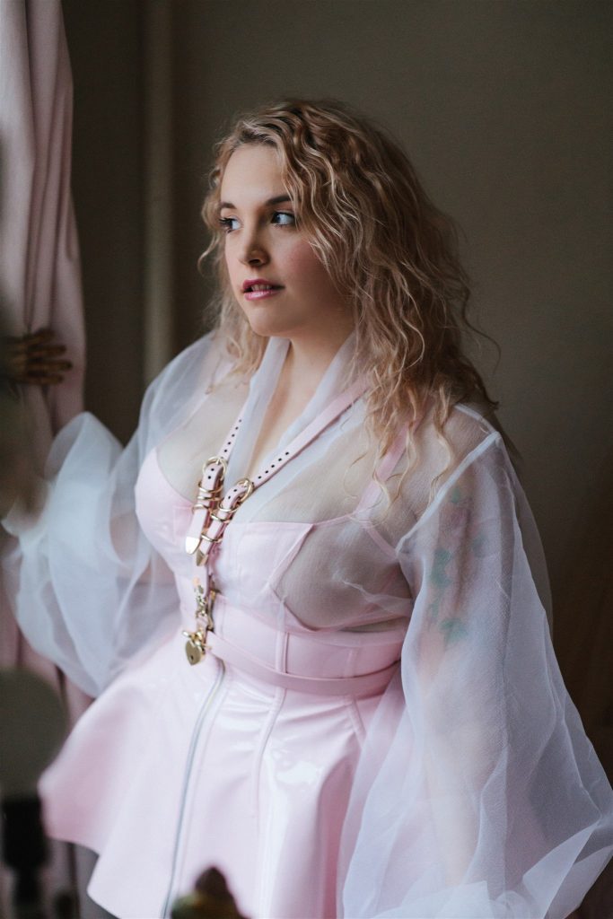 Model standing, wearing light pink PVC corset skirt and Creepyyeha light pink harness.