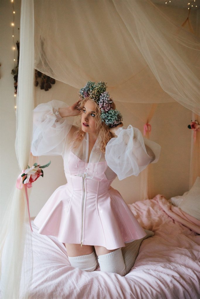 Model kneeling in bed, wearing light pink Artifice custom made PVC corset skirt with high waist and front zipper. 