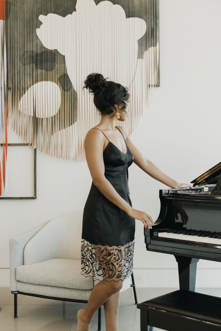 Hadal Lingerie love language dress. Dress has deep hem of black arabic calligraphy on sheer tulle. Model stands next to Piano in front of art work.