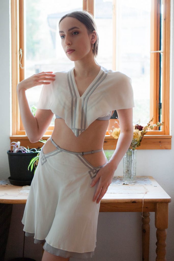 Model in silk lingerie outfit. Pillowbook white and gray silk capelet, bralette and half slip. 