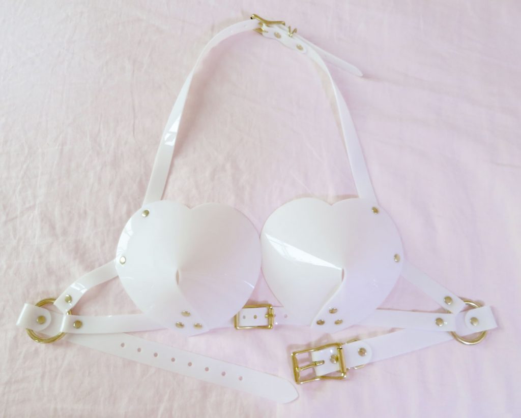 Close up of Apatico, white, PVC bullet bra with heart shaped cups and gold tone buckle straps. laid out on sheet. 