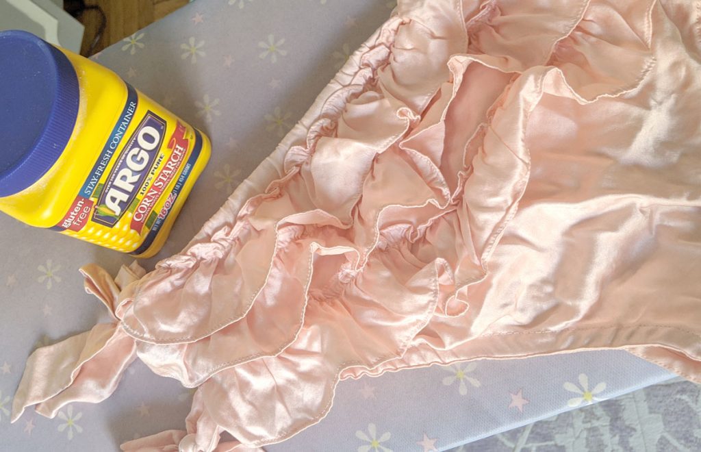 Use cornstarch to remove oil stains from silk lingerie. Photo is of soft pink ruffled silk knickers on an ironing board, with a plastic container of Argo cornstarch next to them.